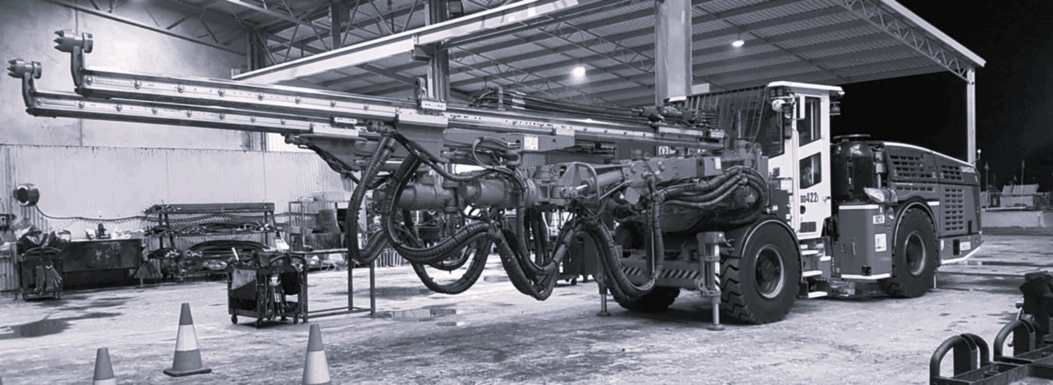 A large, red and white two-boom underground jumbo drill rig used for tunneling and blasting prep in a mine. The long, hydraulic drill booms are extended forward in a maintenance bay
